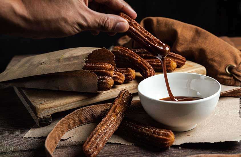 Mão segurando um churros fresco e crocante, coberto generosamente com chocolate, em meio a uma mesa repleta de churros recém-preparados