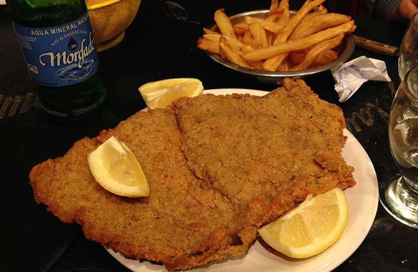 bife à milanesa servido com pedaços de limão em cima de um prato branco, acompanhado por uma porção de batata frita em uma mesa de um dos restaurantes em Bariloche