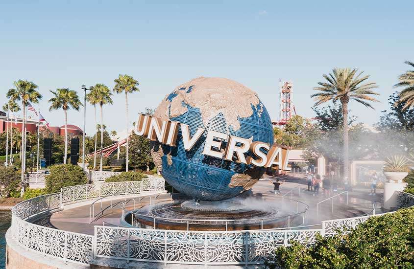 durante o dia, árvores ao redor de grande globo azul com palavra 'universal' ao redor, em entrada de parque da universal studios