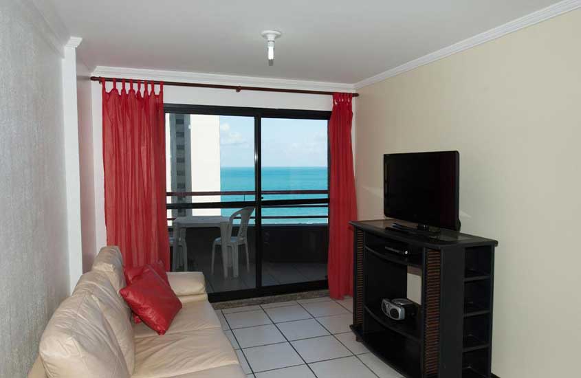 Sofá em frente a uma estante com televisão, em uma espaçosa sala de estar de um apartamento com varanda e vista para o mar