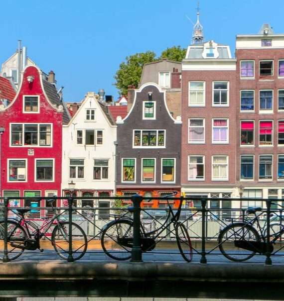 Várias bicicletas estacionadas em uma ponte em Amsterdam durante o dia. Ao fundo, prédios coloridos completam a paisagem.