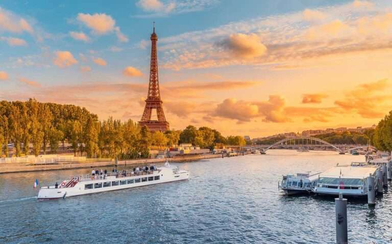 durante um entardecer, vista aérea de barco branco em rio sena, passando em frente a Torre Eiffel