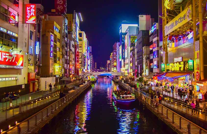 Pessoas caminhando à noite em ruas com letreiros iluminados, à margem de um rio em Osaka, uma das cidades perto de Tokyo.