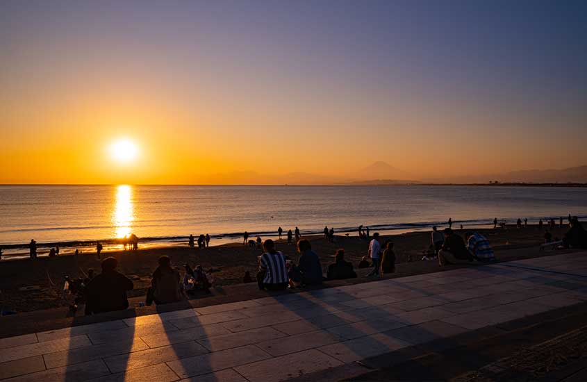Durante um entardecer, diversas pessoas sentadas em grama, em frente ao mar, observando o sol de pôr no horizonte de Enoshima, um dos lugares perto de Tóquio.