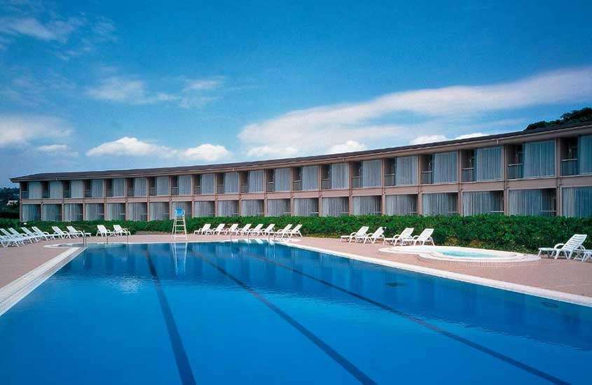 Durante um dia ensolarado, diversas espreguiçadeiras brancas em frente a grande piscina retangular ao ar livre em área de lazer de hotel.