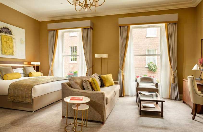 durante o dia, cama de casal, sofá e poltrona em suíte de um dos hotéis em Dublin com janelas amplas e vista para a cidade