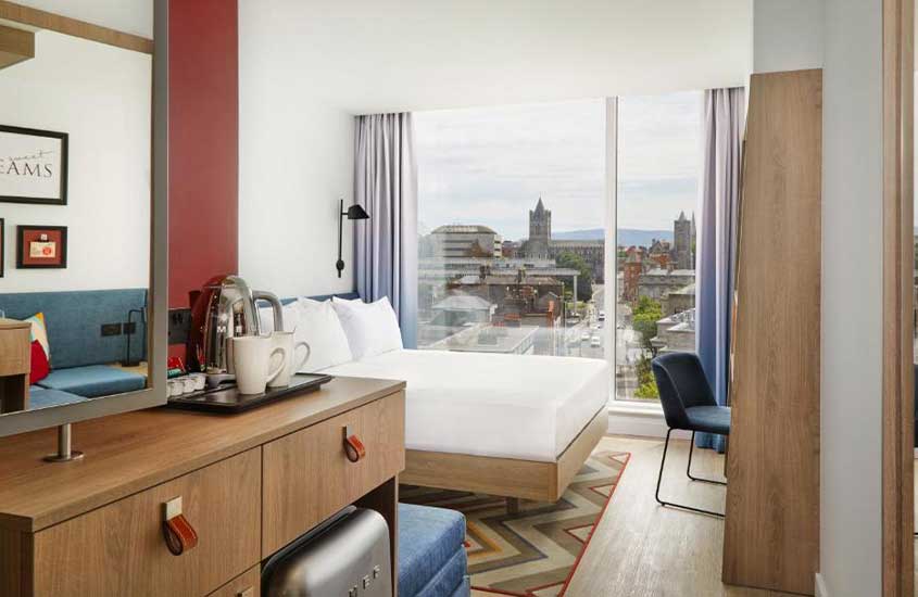 durante um dia nublado, cômoda marrom, cama de casal e cadeira azul em suíte de hotel com janelas do chão ao teto com vista para a cidade