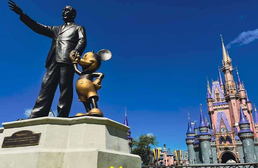 Durante o dia, estátua de Walt Disney de mãos dadas com estátua de personagem Mickey Mouse, e ao fundo um castelo rosa, roxo e azul de parque da Disney