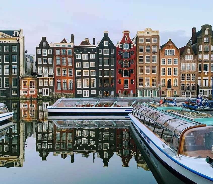 Diversos barcos navegando em um dos canais de Amsterdam durante o dia. Ao fundo, prédios coloridos complementam a paisagem.