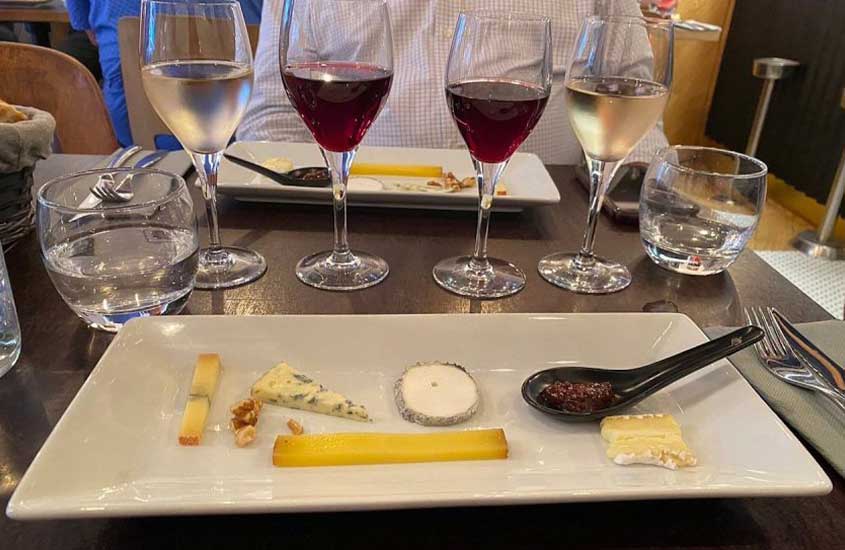 diversos pedaços de queijos servidos em prato branco, junto com taças de vinho, em cima de mesa preta