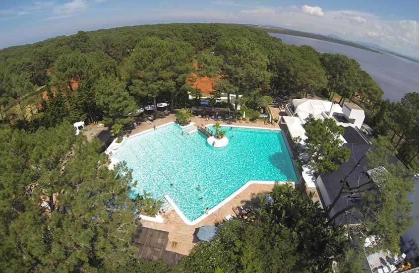 vista aérea, durante um dia ensolarado, de árvores ao redor de piscina ao ar livre em área de lazer de hotel e, ao fundo, mar