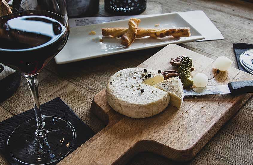 taça de vidro com vidro tinto e pedaço de queijo servido em tábua de madeira em cima de mesa de madeira