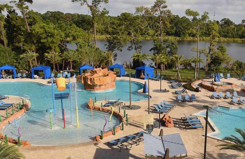 durante o dia, vista aérea de espreguiçadeiras, guarda-sóis e piscinas em complexo de lazer externo de um dos hotéis da Disney em Orlando com vista para um lago.