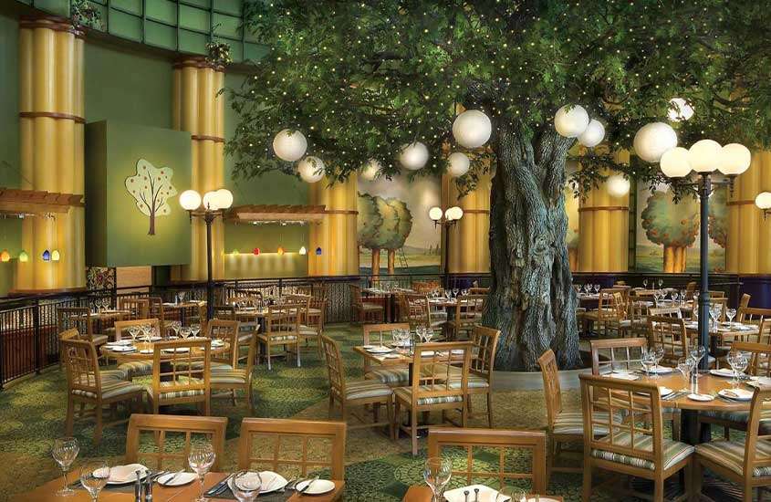 Mesas e cadeiras de madeira em um restaurante de hotel da Disney, decorado com papéis de parede coloridos e uma árvore artificial, criando um ambiente encantador e alegre.