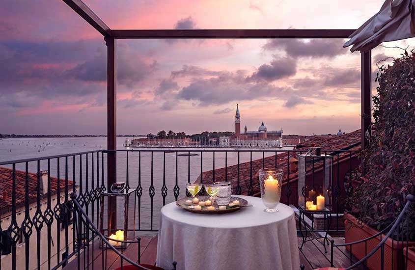 Entardecer em uma varanda de hotel em Veneza, com mesa de jantar decorada com velas e taças de bebidas, e vista dos canais.