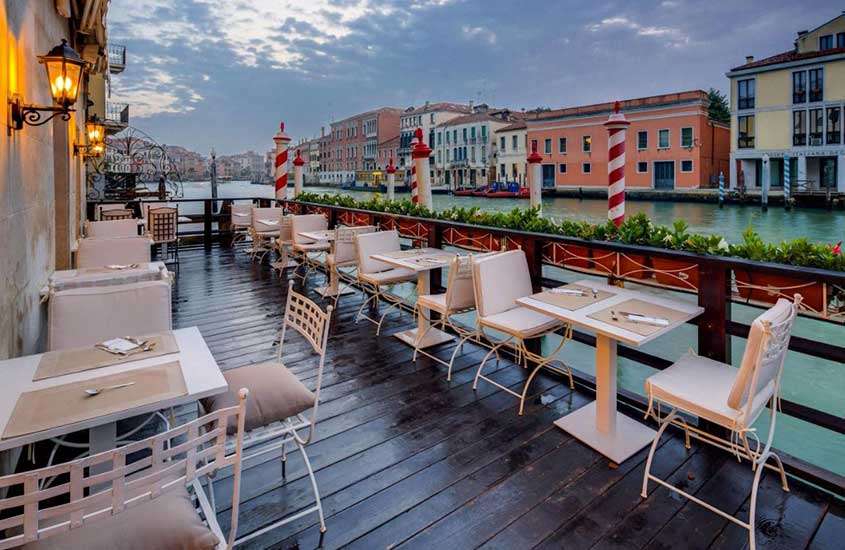 durante um entardecer, mesas e cadeiras em varanda de hotel em Veneza com vista para o canal.