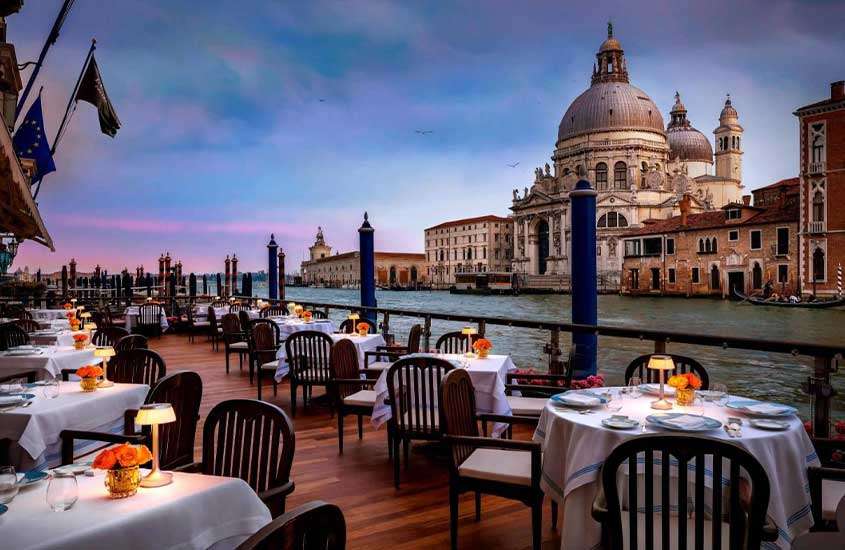 durante um entardecer, mesas e cadeiras em varanda de hotel em Veneza com vista para o canal.