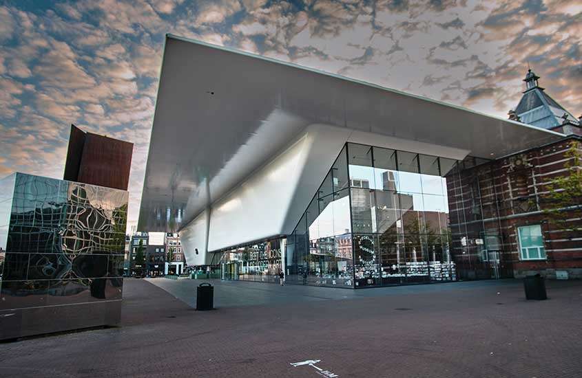 durante o dia, prédio com paredes de vidro e grande telhado branco, onde funciona Stedelijk, um dos principais museus de amsterdam