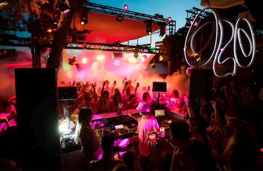 Durante o anoitecer, balada em Ibiza com pessoas, dj, fumaça, luzes coloridas e árvores ao redor