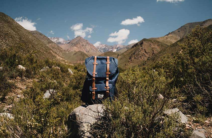 Em um dia ensolarado, uma mochila azul colocada em cima de uma pedra, em uma trilha com vista panorâmica das montanhas.