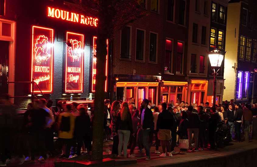 Durante a noite, pessoas reunidas na porta de uma fachada de vitrine iluminada por luzes vermelhas no Red Light District