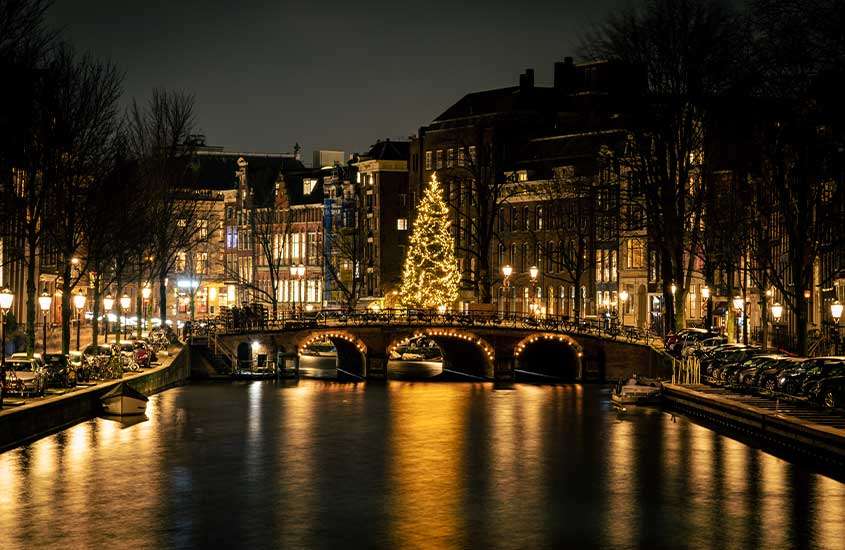 Durante a noite, vista panorâmica de ponte sobre lago, iluminada com luzes. Ao fundo, árvores de natal e prédios