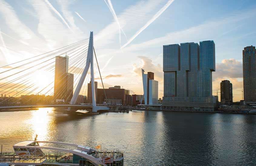 Durante um entardecer, vista panorâmica de ponte sobre rio em Rotterdam. Ao fundo, construções modernas
