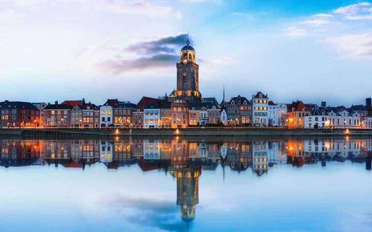 Durante entardecer, vista panorâmica de construções iluminadas às margens de rio, em uma das cidades da Holanda