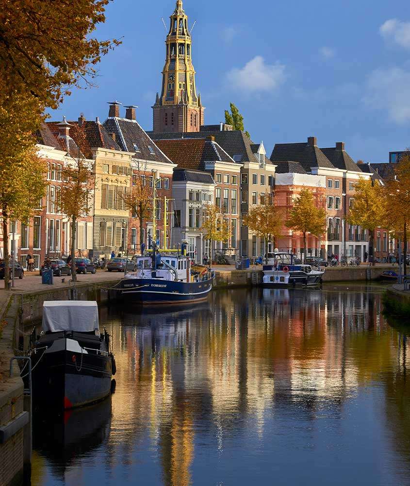 durante dia ensolarado, prédios coloridos, igreja e árvores alaranjadas refletidas em rio em uma das cidades da holanda