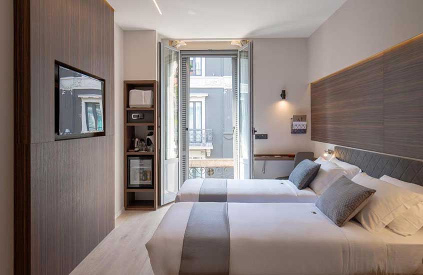 Durante o dia, quarto de hotel em Milão mobiliado com TV e duas camas de solteiro. Ao fundo, varanda com vista para a cidade.