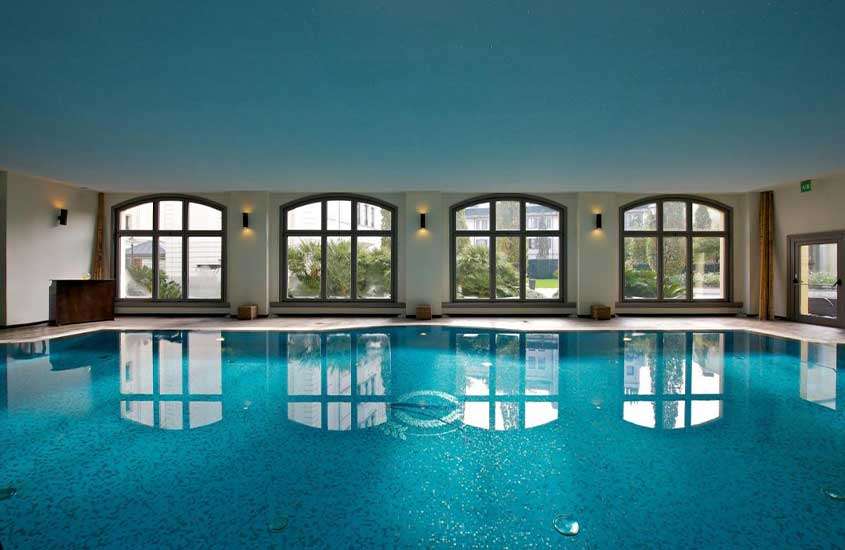Durante o dia, grande piscina retangular em área de lazer de hotel coberta com diversas janelas.