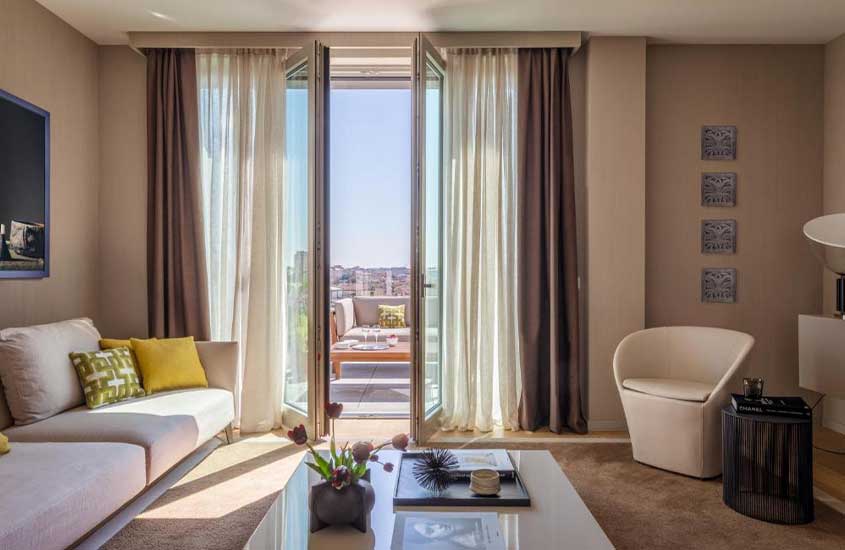 sala de estar de suíte em hotel em Milão, mobiliada com sofá, poltrona e mesa de centra. Ao fundo, varanda com sofá e vista da cidade.