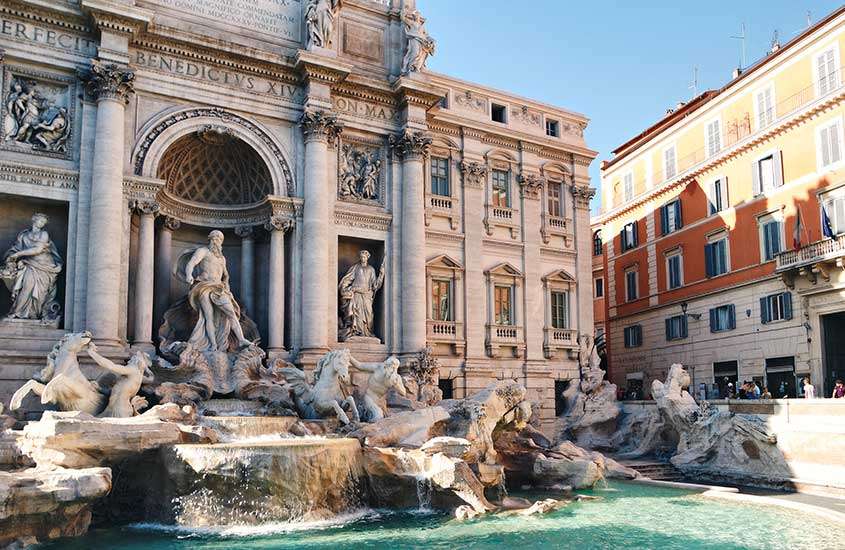 Durante o dia, vista da Fontana di Trevi, fonte com várias esculturas em mármore de deuses e águas azuis, rodeada por construções históricas.
