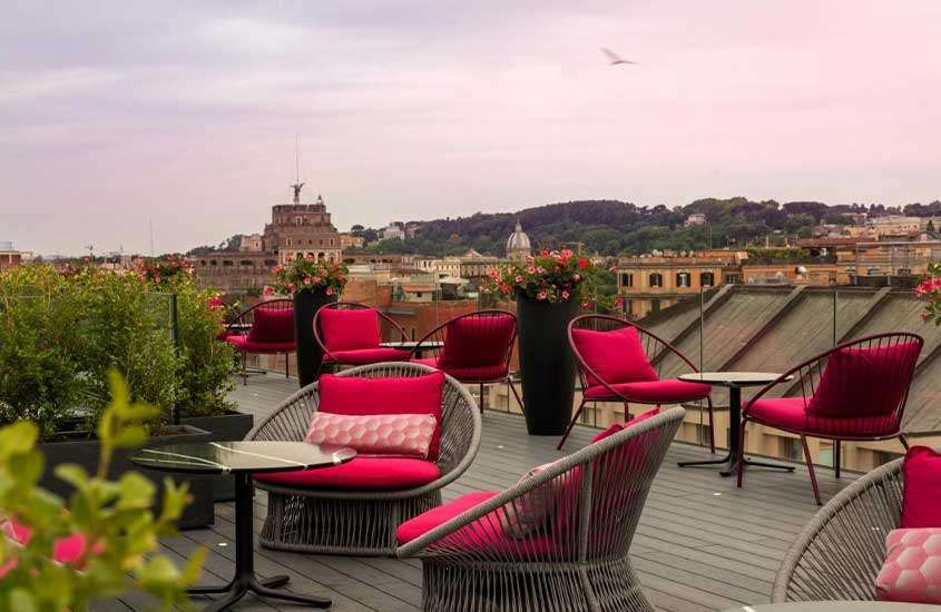 Ao entardecer, mesas e cadeiras acolchoadas, em terraço de hotel em Roma com vista da cidade.