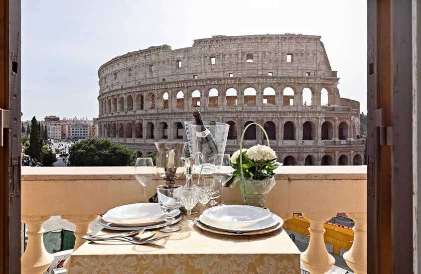 Em um dia ensolarado, mesa posta com louça, taças de cristal, champagne e flores brancas em varanda de suíte de hotel em Roma com vista para o Coliseu