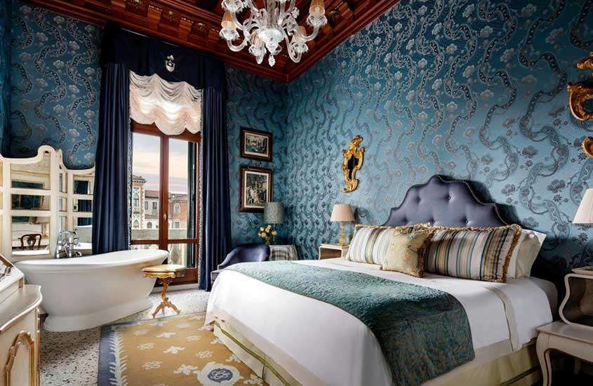 Durante o dia, suíte de hotel em Veneza mobiliada com uma banheira, espelho, cama de casal e poltrona, proporcionando conforto e luxo aos hóspedes.