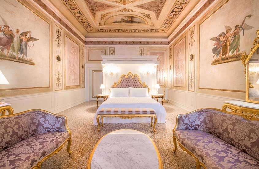 Suíte de hotel em Veneza luxuosa, com belas pinturas nas paredes e teto, e equipada com sofás e cama de casal de madeira e detalhes dourados.