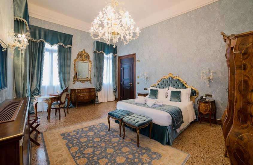 Quarto de um dos hotéis em Veneza, elegantemente mobiliado com guarda-roupa, mesas, cadeiras e cama de madeira, em tons suaves de azul.