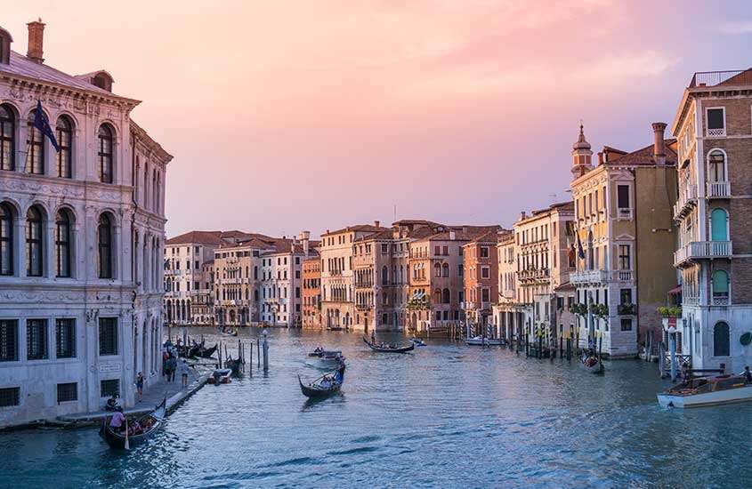 Durante entardecer, vista aérea de gondolas em um dos canais de Veneza, rodeado por prédios coloridos.