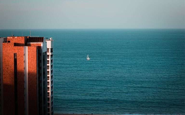 Em dia de sol, vista aérea panorâmica de mar, barco a vela e um pedaço da prédio à esquerda