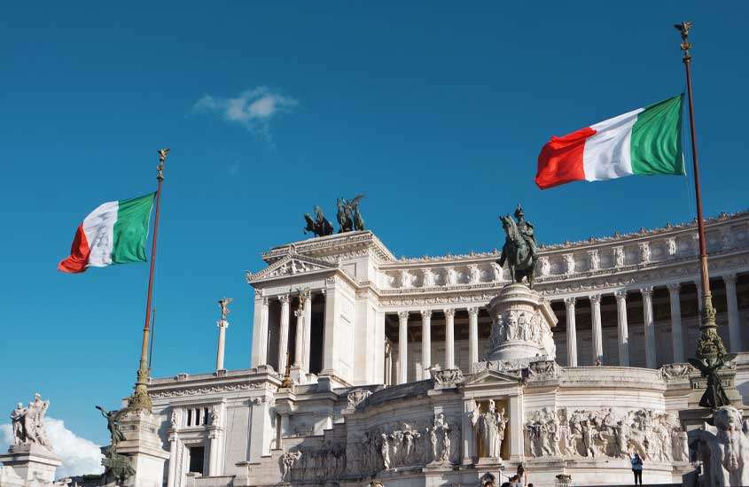 Fórum Romano, esculturas de mármore e cobre iluminadas pelo sol da tarde, com o céu azul ao fundo e as bandeiras da Itália tremulando
