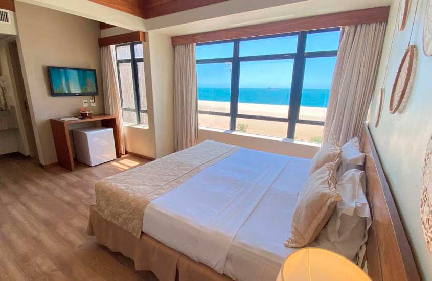 Em dia de sol, quarto com cama de casal, luminárias, televisão, frigobar e grandes janelas com vista para o mar
