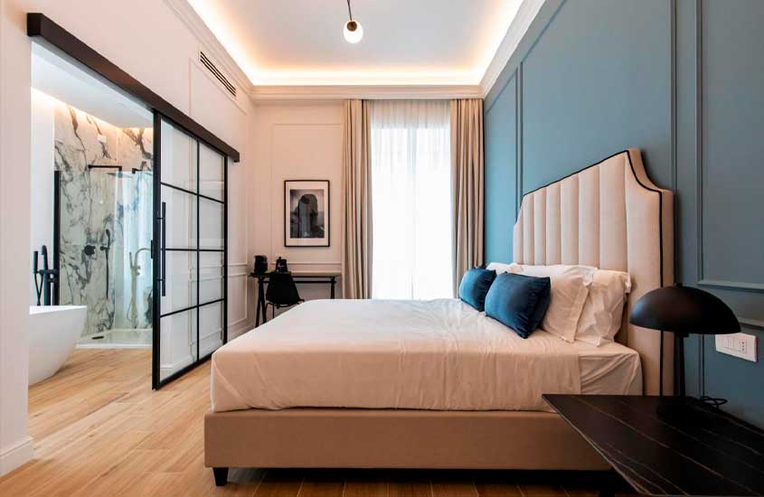 quarto de hotel em Nápoles em tons neutros com cama de casal, estação de trabalho, quadro, cortina e lustre simples