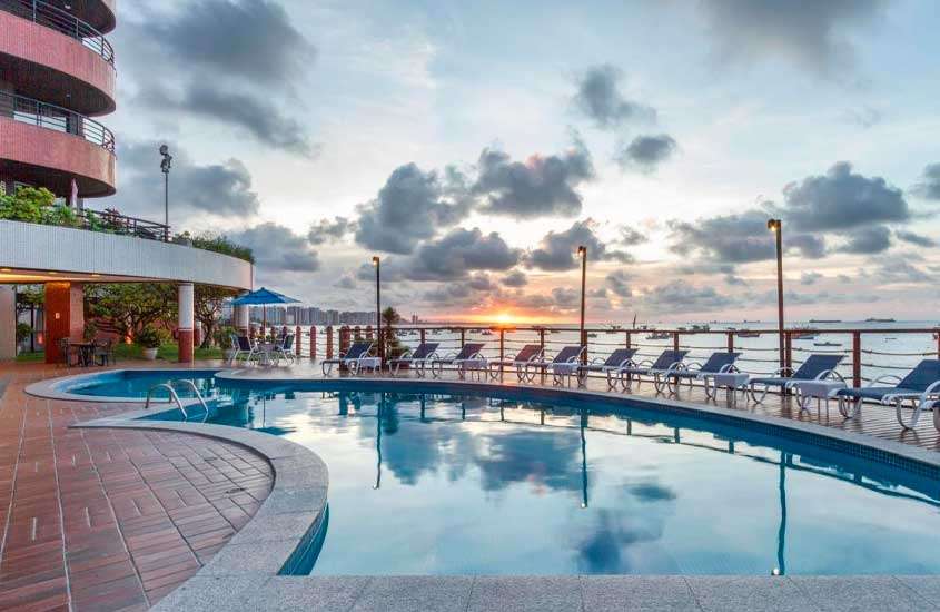 Durante o pôr do sol, área de lazer de hotel com piscina externa, espreguiçadeiras, guarda-sóis, quiosque e vista para o mar