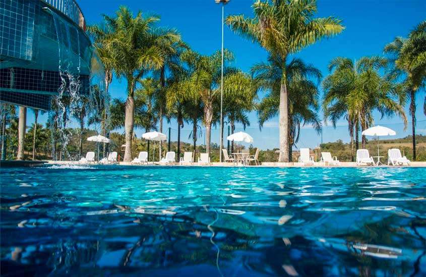 durante o dia, piscina ao ar livre rodeada por palmeiras, espreguiçadeiras com guarda-sol. Ao fundo, vista das montanhas