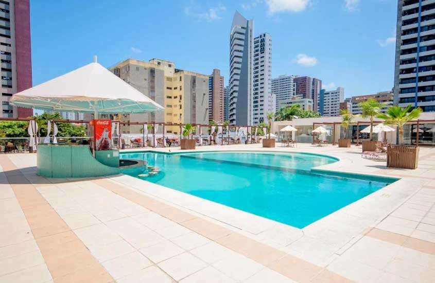 Em dia de sol, piscina em área de lazer externa de hotel com bar molhado guarda-sóis, mesas, cadeiras, vasos de planta e vista da cidade