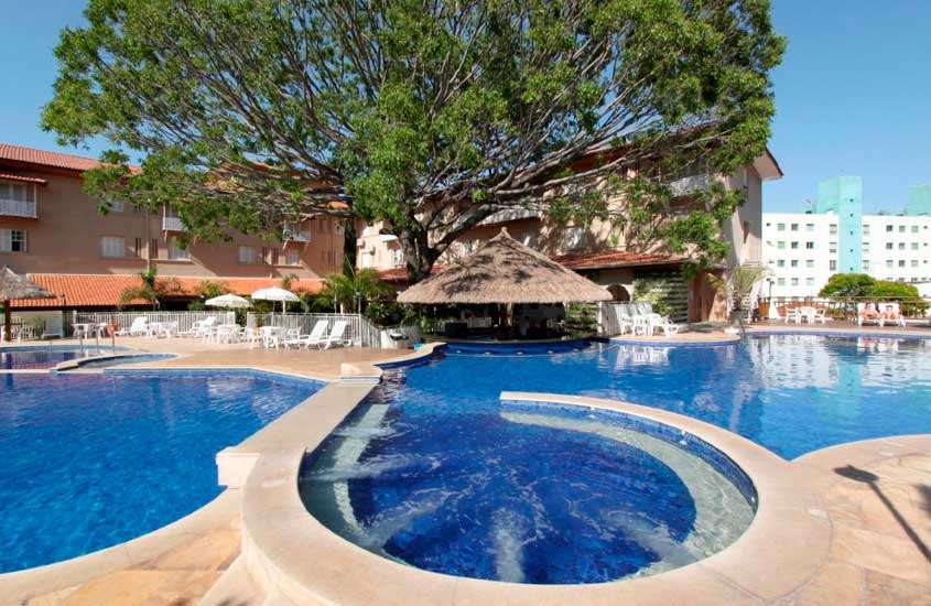 durante um dia ensolarado, área de lazer de hotel em aguas de lindoia com piscina, bar molhado, espreguiçadeiras brancas e grande árvore em frente