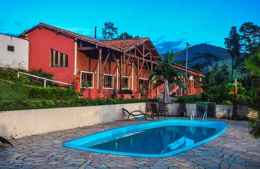 Durante o entardecer, vista de piscina externa de um hotel fazenda no Sul de Minas com espreguiçadeira, mesa, cadeiras, guarda-sol e hotel ao fundo