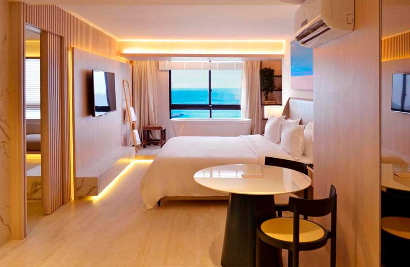 Em dia de sol, quarto de hotel com mesa e cadeiras altas, cama de casal, televisão, ar condicionado, toalheiro e janela com vista para o mar