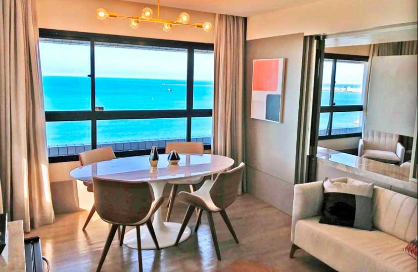 Em dia de sol, sala de suíte de hotel com mesa, cadeiras, sofá, espelho, janela grande com cortina e vista para o mar